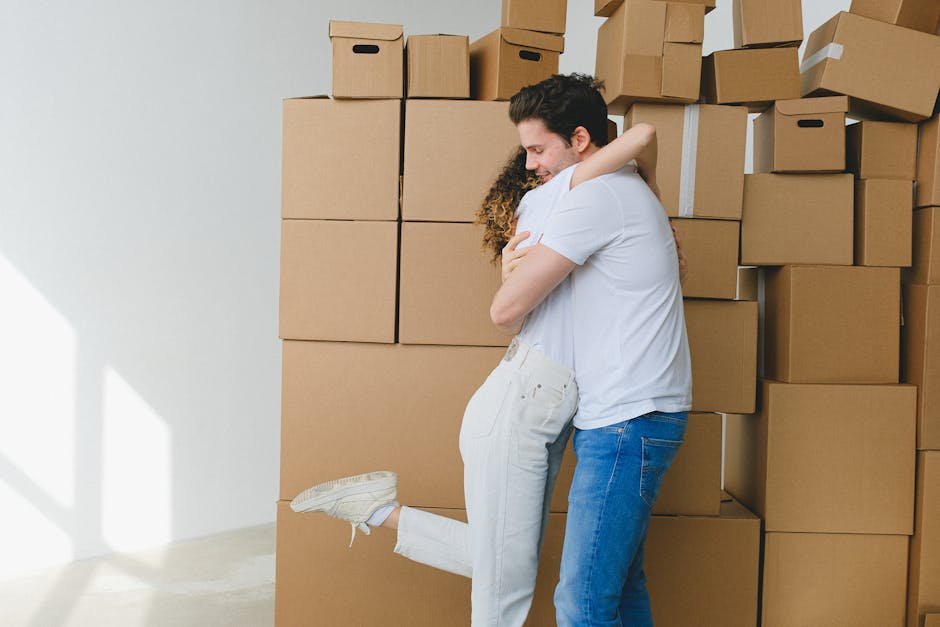 Inside a well-lit room with light-colored walls and a large window with blue curtains, two individuals are preparing for a home relocation process, representing a furniture transport and packing activ